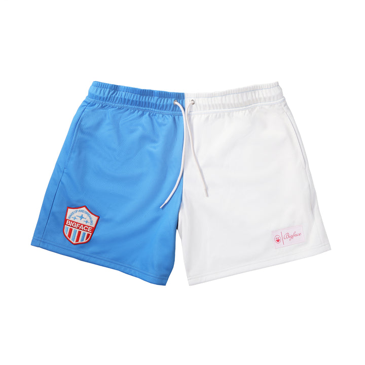 A pair of shorts, one leg blue and one white, laid flat. The shorts have a drawcord and a zigzag stitch patch on the front, with a visible brand tag on the front.